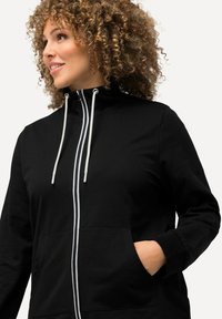 Giacca nera con zip e colletto alto, due tasche frontali e dettagli con cordini bianchi. Realizzata in un tessuto morbido e texture.
