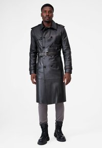 Black leather trench coat with double-breasted buttons, belted waist, sharp collar, and shoulder epaulettes, paired with gray pants and black boots.