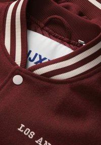 La veste varsity bordeaux présente un col côtelé avec des rayures crème, une fermeture à boutons et un texte brodé qui indique "LOS ANGELES."