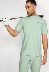 Ανοιχτό πράσινο γκολφ πουκάμισο με ψηλό γιακά, διακοσμημένο με διακριτικό μοτίβο και λογότυπο στο στήθος. Συνδυάζεται με παντελόνια στο ίδιο χρώμα.