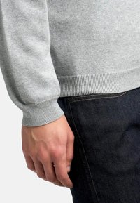 Suéter gris claro con puños acanalados, llevado sobre jeans de mezclilla oscuros. Primer plano muestra una mano descansando sobre el bolsillo lateral de los jeans.