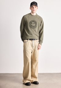 Olivovo zelená mikina s kruhovým logom, béžové rifle s širokými nohavicami, čierny opasok a lesklé čierne topánky, doplnené čiernou čiapkou.