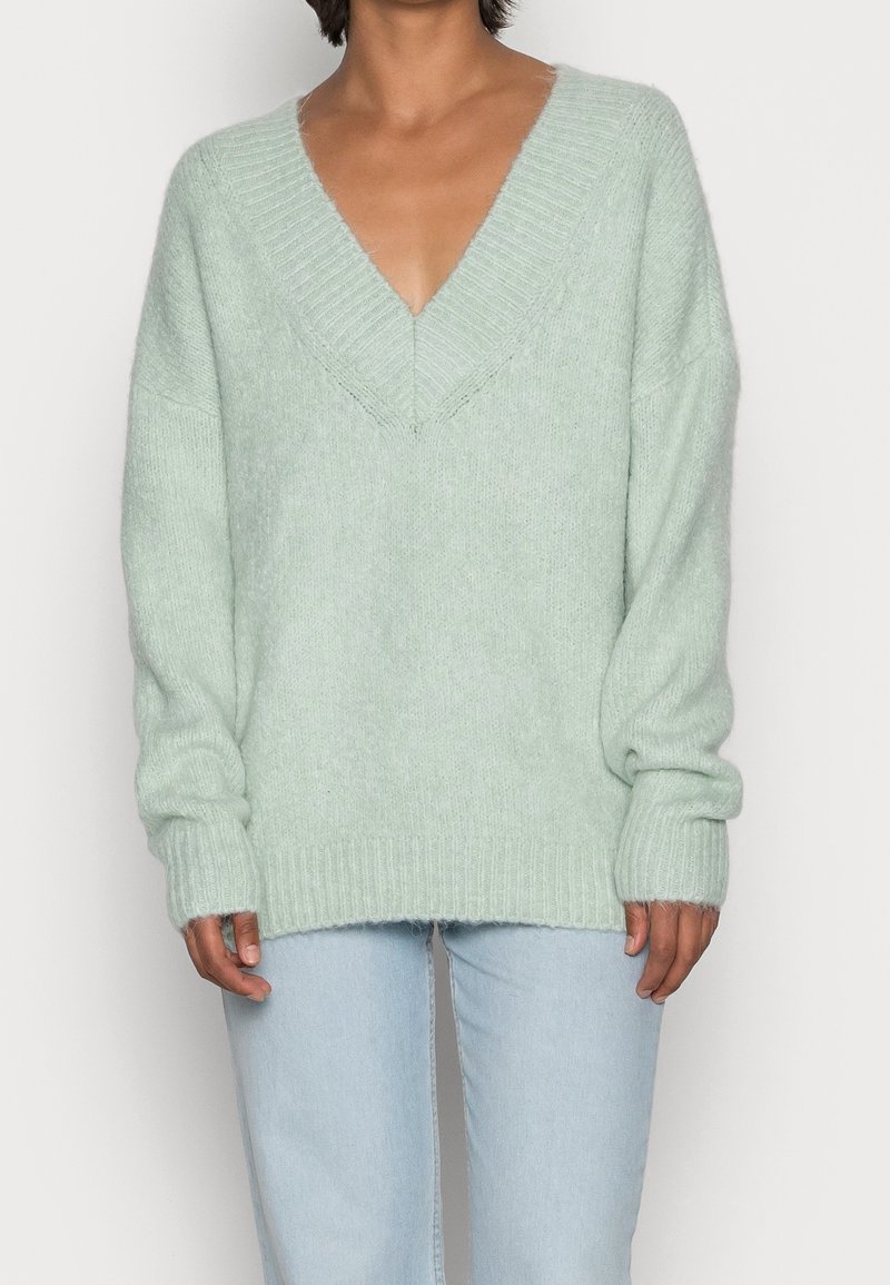 Personne portant un pull à col en V duveteux vert clair à manches longues et un jean bleu clair sur un fond uni.
