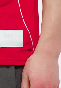 Närbild på en persons arm och hand intill en röd tröja med en vit etikett som visar numret "1892".