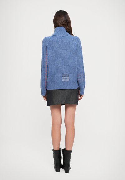 Maglione blu lavorato a maglia con colletto alto, caratterizzato da un motivo a quadretti sul retro e dettagli rossi lungo le maniche. Indossato su una gonna scura.