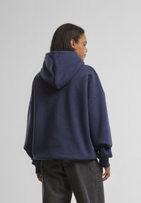 Mørkeblå oversized hettegenser med snøring i hetta, ribbestrikkede mansjetter og glatt tekstur, kombinert med mørk grå jeans.