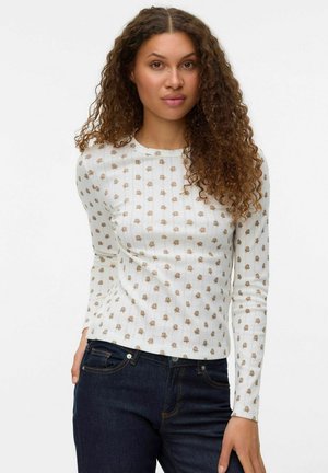 Witte langetermijn shirt met een geribbelde textuur, voorzien van een herhalend gouden bomenpatroon. Gecombineerd met donkerblauwe jeans.