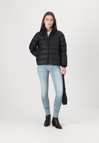 Svart puffjakke med høy krage, glidelås og to sidelommer, kombinert med lyseblå jeans og svarte ankelstøvler.