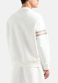 Weißes Sweatshirt mit Rundhalsausschnitt, gerippten Bündchen und zwei horizontalen beigen Streifen auf dem Ärmel. Glatte Stoffoberfläche.