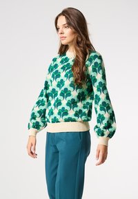 Grüner und cremefarbener Pullover mit einem Blättermuster, Rundhalsausschnitt und puffierten Ärmeln. Rippenbündchen und Saum. Kombiniert mit türkisfarbenen Hosen.