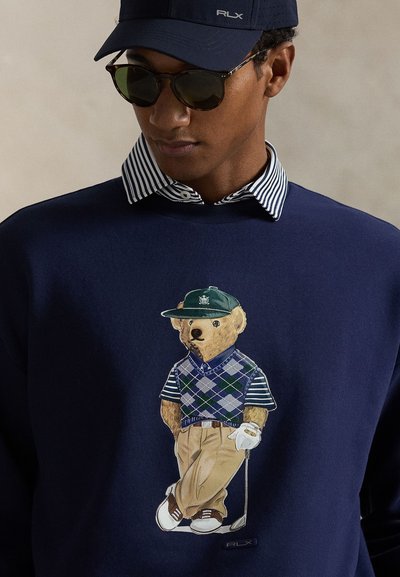 Μπλε ναυτικό φούτερ με γραφικό αρκούδας που φορά πράσινο καπέλο, γιλέκο αργκλάι, και χακί παντελόνι, με γυαλιά ηλίου και λεπτομέρεια γκολφ κλαμπ.