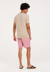 Camiseta beige de mangas cortas, pantalones cortos a rayas rosas, sandalias marrones. Costuras y un bolsillo visibles en los pantalones cortos. Modelo de espaldas a la cámara.