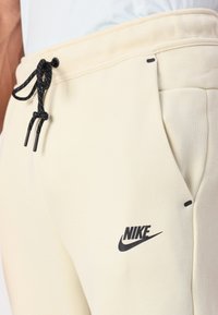 Cremfarbene Nike-Sweatpants mit schwarzem Kordelzug, Seitentaschen und Logo. Glatte Textur mit maßgeschneidertem Schnitt und minimalistischem Design.