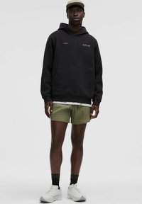 Mann steht und trägt einen schwarzen Kapuzenpullover, olivgrüne strukturierte Shorts, schwarze Socken, weiße Sneakers und eine beige Mütze vor einem schlichten Hintergrund.