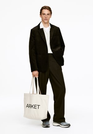 Schwarzer Cord-Blazer mit zwei Taschen, kombiniert mit schwarzen Hosen und grauen Sneakers. Hält eine beigen Tasche mit dem Aufdruck "ARKET" in Schwarz.