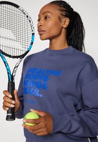 Marinblå sweatshirt med blå text som säger "HÄLSA & KROPP & SINNE & SJÄL." Tennisracket och gul boll hålls i händerna, uppvisat mot en vit bakgrund.