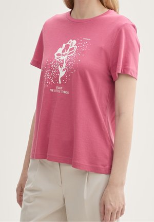 Vrouw draagt een roze T-shirt met korte mouwen met een witte bloemafbeelding en tekst "Enjoy the little things", gecombineerd met beige broek.
