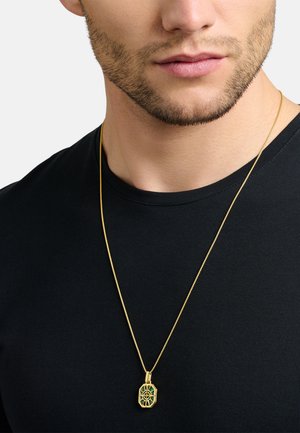 Goldene Halskette mit einer grünen, geometrischen Anhänger mit filigranen Ausschnitten, vor einem schwarzen T-Shirt-Hintergrund.