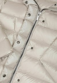 Chaqueta tipo plumas en beige claro, con diseño acolchado, cremallera plateada y botones a presión. Textura brillante y suave con una silueta estilizada.
