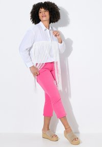 Bílá košile s knoflíky a detaily fringing, růžové upnuté kalhoty a béžové platformové sandály. Na bílém pozadí, s kudrnatými vlasy.