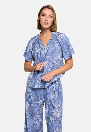 Mujer vistiendo una blusa de estampado floral azul y pantalones anchos a juego, sosteniendo un collar largo con borlas azules sobre un fondo blanco.