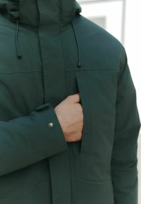 Dark green, insulated jacket with a hood. Features a visible chest pocket and adjustable drawstrings. Material has a subtle check pattern.