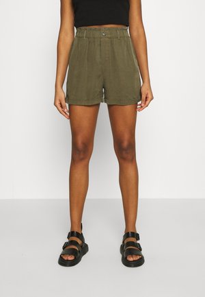 Kvinde iført olivengrøn shorts med høj talje, sort cropped top og sorte sandaler med dobbelte remme, står foran en ensfarvet baggrund.