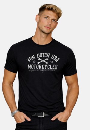 Jeune homme aux cheveux blonds courts portant un t-shirt noir avec l'inscription « Von Dutch USA Motorcycles » et un motif de clés croisées.
