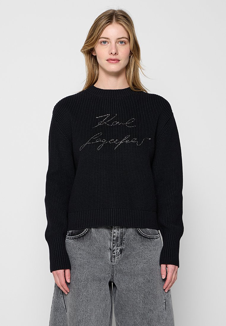 Pull en tricot noir avec une texture côtelée, présentant un design court et des détails en script métallique à l'avant. Manches longues présentes.