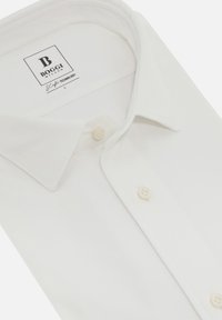 Bílá košile s knoflíky vyrobená z texturované látky. Má klasický límec a dva přední knoflíky. Na etiketě jsou uvedeny informace o značce a technologii.