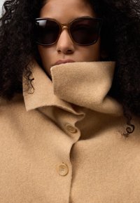 Cappotto di lana beige con colletto alto e tre bottoni. Il modello indossa occhiali da sole scuri. Il tessuto appare morbido con una texture liscia.