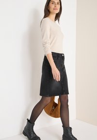 Gonna di denim nera con vita aderente, lunghezza fino al ginocchio, abbinata a un maglione beige chiaro e stivali neri con lacci. Borsa marrone a tracolla.