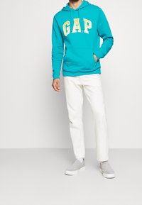 Sudadera con capucha turquesa con logo amarillo "GAP", bolsillo delantero, combinada con pantalones blancos y zapatos tipo slip-on grises, sobre un fondo liso.