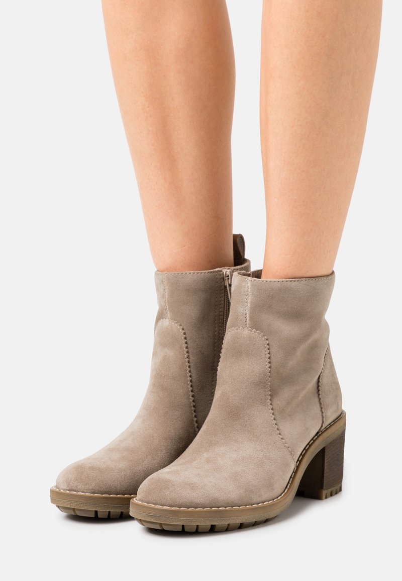 Bottes en daim taupe montantes à la cheville avec fermetures éclair latérales et talons carrés, portées sur des jambes nues, présentées sur un fond clair.