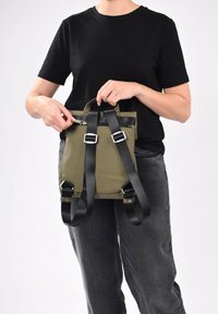 Mochila de color verde oliva hecha de tela con correas ajustables negras, que cuenta con un asa superior, un bolsillo delantero con cremallera y hebillas metálicas.