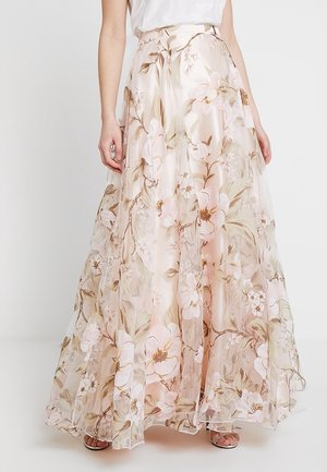 Jupe maxi fluide avec un imprimé floral rose pâle et beige, portée avec un top blanc et des chaussures ouvertes couvrant presque entièrement les pieds.