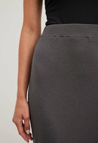Šedá žebrovaná sukně s přiléhavým střihem, elastickým pasem a hladkým povrchem. Detailní záběr s rukou položenou na boku.