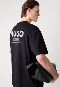 Melna kokvilnas T-kreklu ar izteiksmīgu balto "HUGO" logotipu uz aizmugures. Modelis tur melnu ādas somu, parādot ikdienišķu stilu.