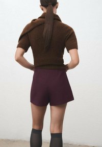 Donna con i capelli lunghi raccolti in una coda di cavallo, che indossa un maglione corto marrone a maniche corte, pantaloni corti borgogna e calze nere sopra il ginocchio, vista di spalle.