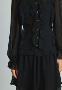 Blusa nera con maniche lunghe trasparenti, dettaglio frontale arricciato e bottoni argentati. Presenta una texture delicata e stratificata con una vita aderente.