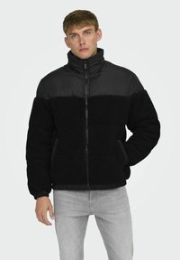 Chaqueta negra de cremallera con un cuerpo de forro polar texturizado y paneles acolchados lisos. Cuello alto y puños elásticos, combinada con jeans grises.