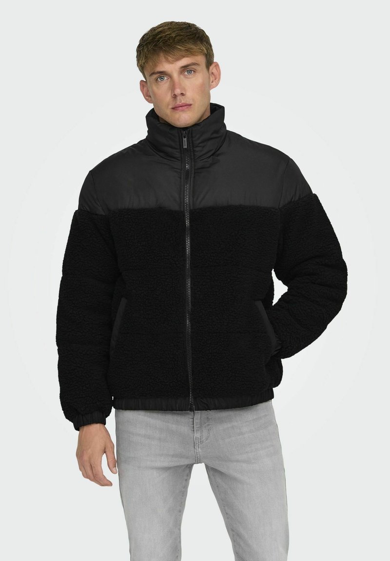 Chaqueta negra de cremallera con un cuerpo de forro polar texturizado y paneles acolchados lisos. Cuello alto y puños elásticos, combinada con jeans grises.