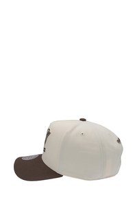 Cream cap with a brown bill, featuring an embroidered logo on the front and stitching details on the crown. Structured design with metal eyelets.