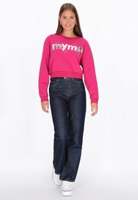 Fukszia színű rövidített pulóver "mymo" grafikával, sötétkék, magas derekú farmernadrággal és fehér tornacipővel, egyszínű háttér előtt állva.