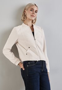 Giacca bomber beige chiaro con chiusura a zip, polsini a coste e due tasche laterali. Abbinata a pantaloni jeans scuri. Tessuto dalla superficie liscia.