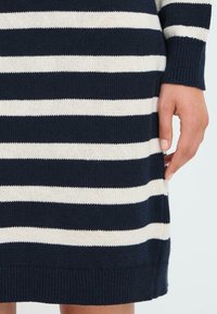 Strikkegenser med marineblå og kremfargede horisontale striper, ribbestrikkede mansjetter og kant. Nærbilde av den nedre delen og hånddetaljer.