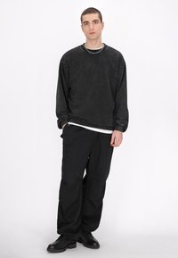 Schwarzer Langarm-Sweatshirt mit texturierter Oberfläche, kombiniert mit lockeren schwarzen Hosen und schwarzen Schuhen. Als Zubehör trägt die Person eine silberne Halskette.