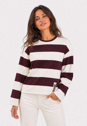 Frau mit welligem braunem Haar, die ein langärmliges Shirt mit breiten burgunderroten und weißen horizontalen Streifen und helle Hosen trägt.