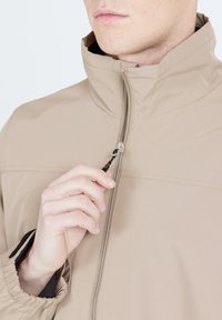 Beige zipjacka med hög krage; texturerat tyg; har en dragsko vid dragkedjan; elastiska ärmslut.