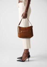 Geanta de umăr din suede ciocolatiu, cu o curea texturată și accesorii aurii, având un accent din lanț. Purpă cu pantaloni scurți crem și tocuri negre.